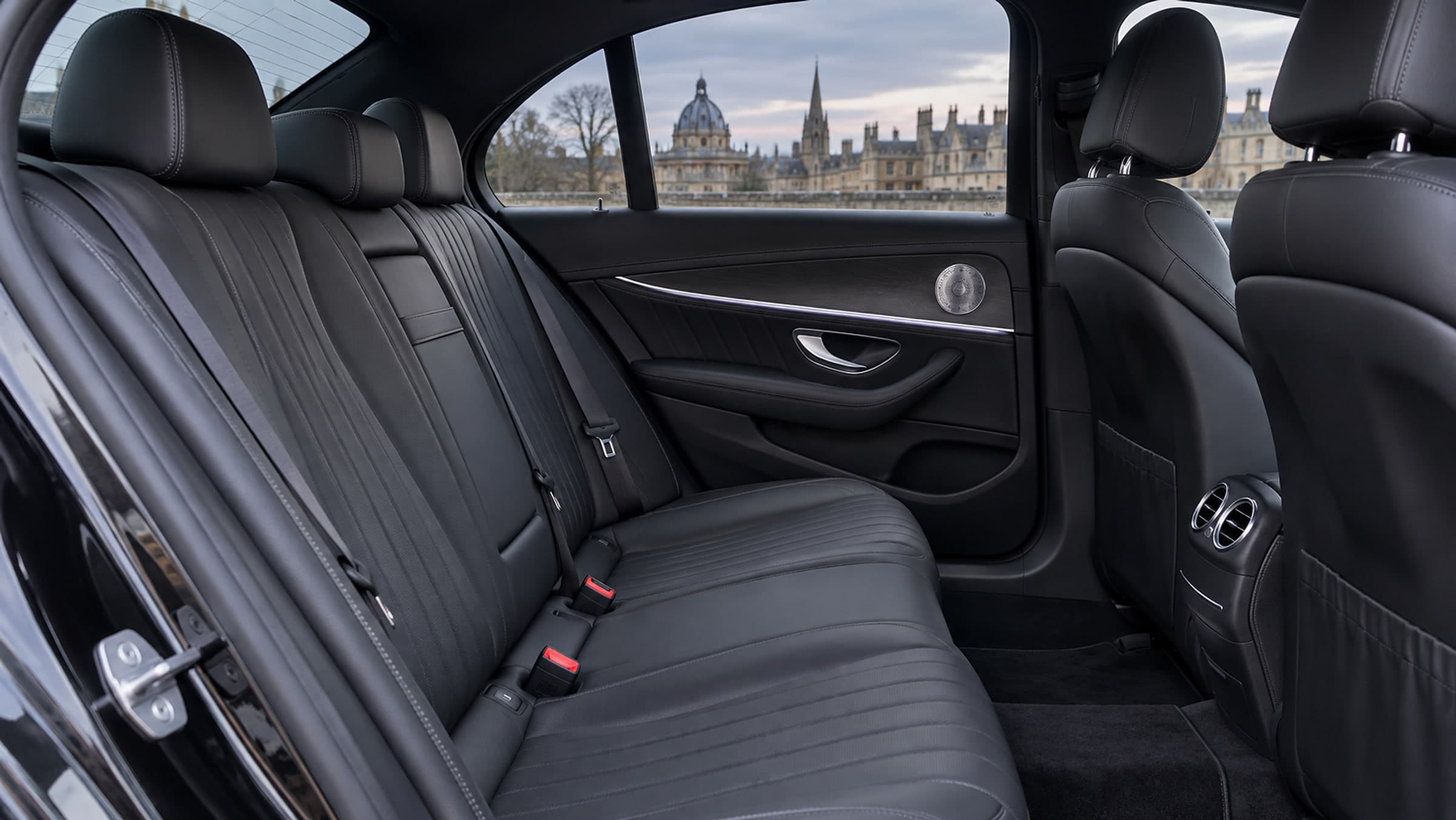 The rear seats of a Mercedes E-Class: a black leather bench, wood and soft ambient light on the door, and Oxford through the window