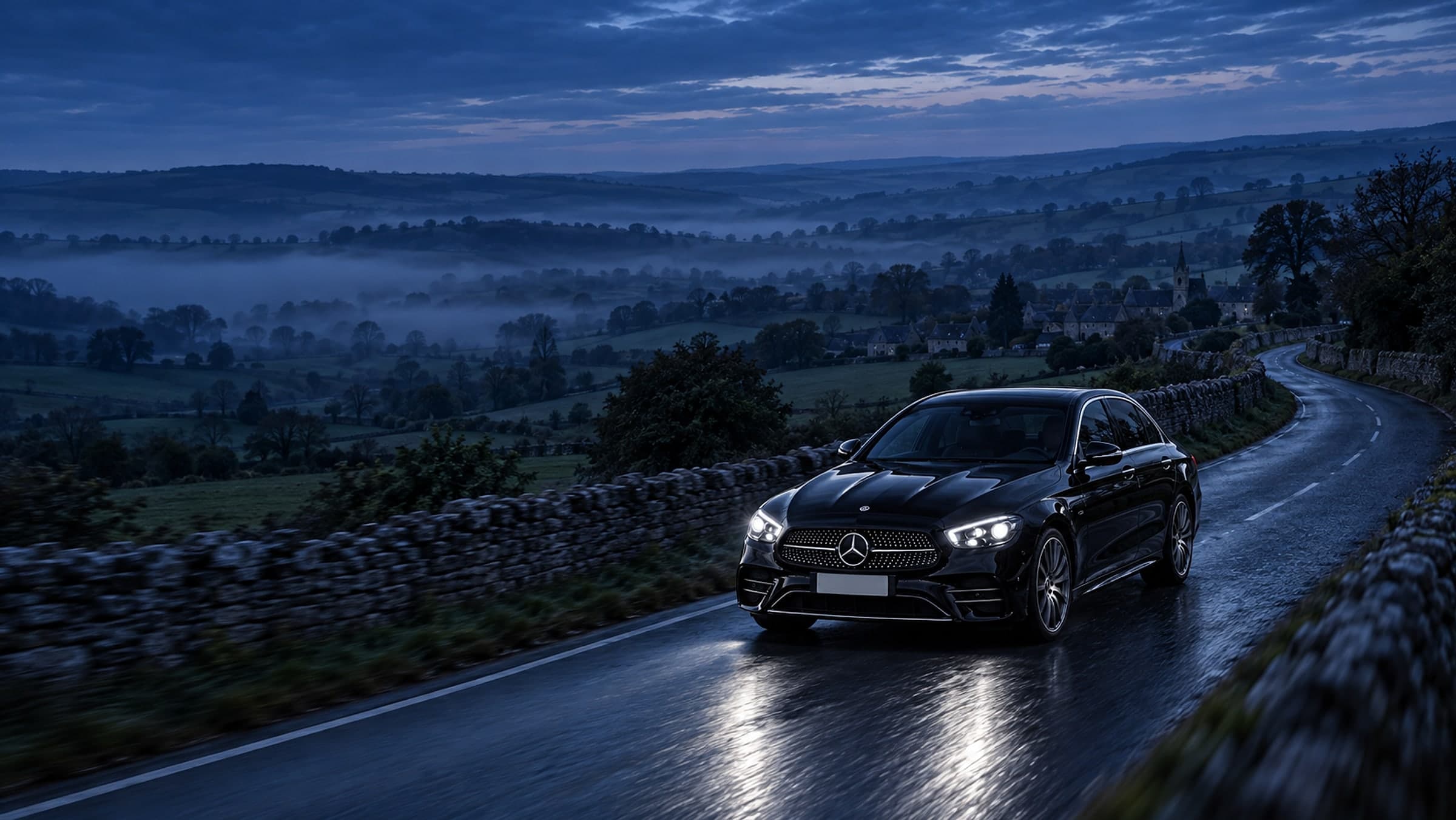 A black Mercedes E-Class Saloon on a winding Cotswolds road at blue hour, mist in the valley